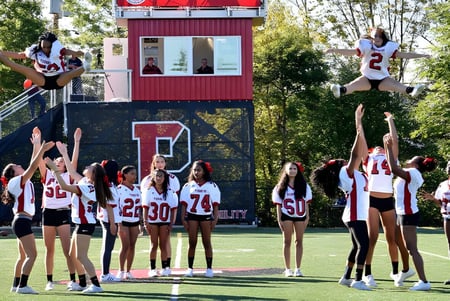 Estudiantes de The Pennington School realizan acrobacias de animadoras en un campo deportivo frente a un edificio rojo y blanco.