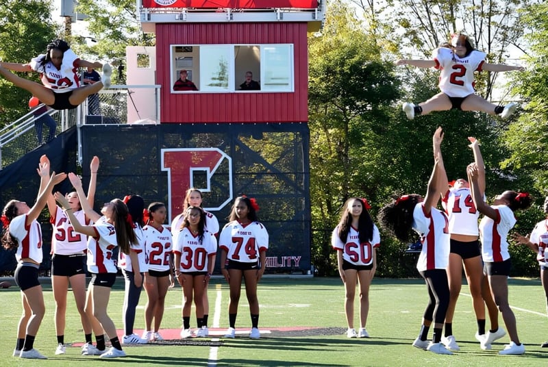 Estudiantes de The Pennington School realizan acrobacias de animadoras en un campo deportivo frente a un edificio rojo y blanco.