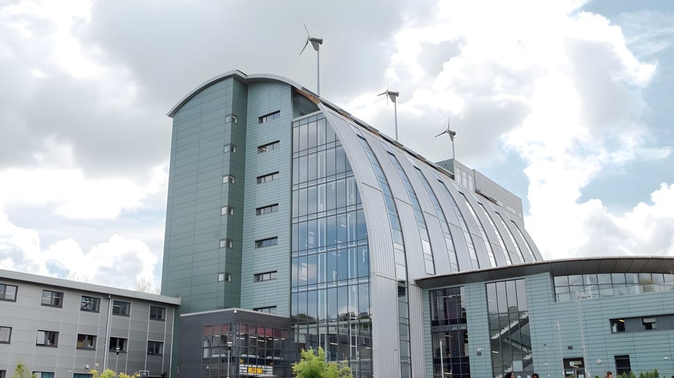 El moderno edificio del The Sheffield College con fachada de vidrio y aerogeneradores en el techo bajo un cielo nublado.
