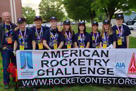 Estudiantes de The Vanguard School posan frente a un banner de la American Rocketry Challenge con árboles y edificios al fondo.