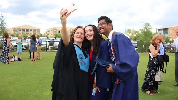 Tres graduados de The Village School se toman un selfie en el campus con edificios de fondo.