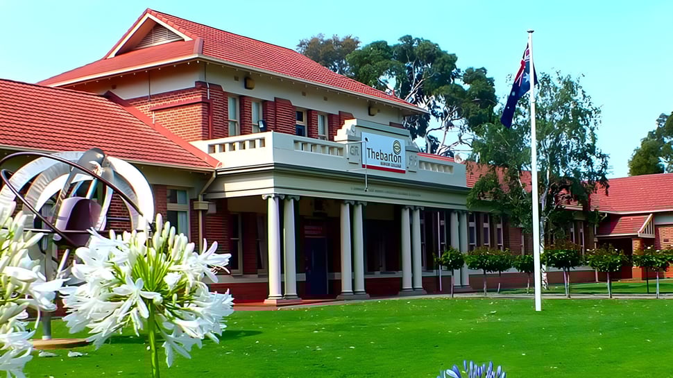 El edificio de varios pisos del Thebarton Senior College con techo rojo y columnas se encuentra en un prado con árboles y flores en primer plano.