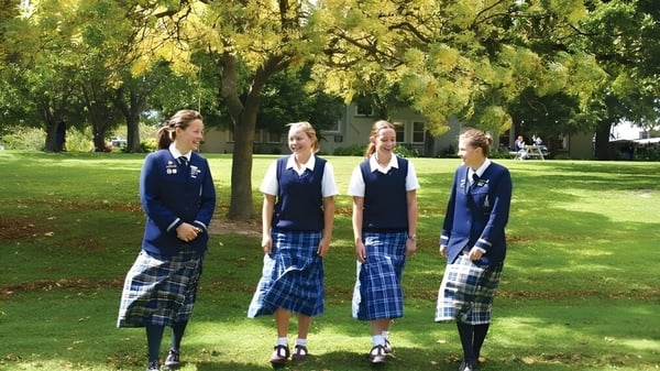 Cuatro alumnas de la Timaru Girls School están en un césped en un parque con árboles de fondo.