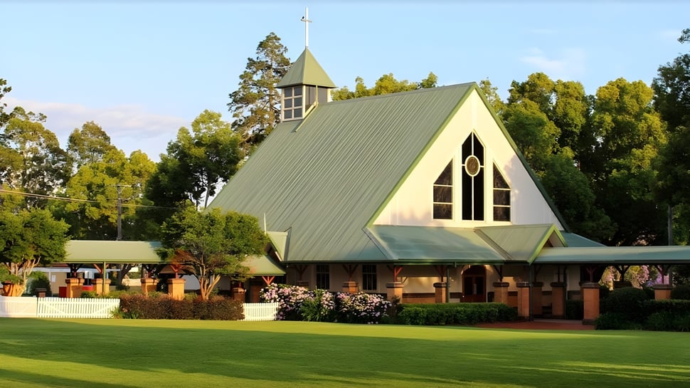 Una iglesia con techo puntiagudo y torre se encuentra en el terreno de la Toowoomba Anglican School rodeada de un paisaje verde y árboles.