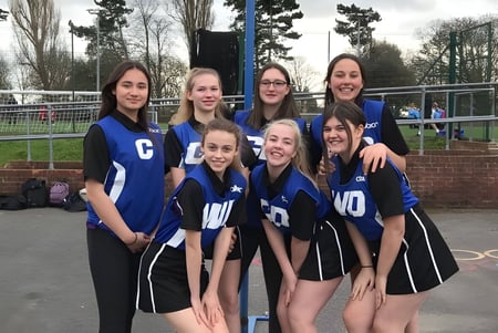 Estudiantes de la Trafalgar School posan juntas en el campo deportivo con árboles y una valla de fondo.
