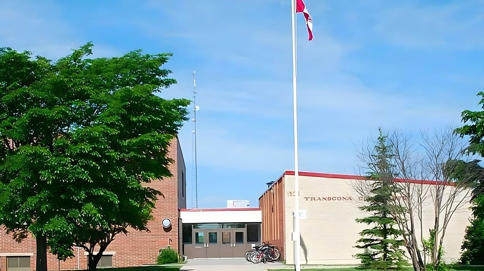 La bandera en el asta ondea frente al edificio de ladrillo de la Transcona Collegiate y está rodeada de árboles verdes.
