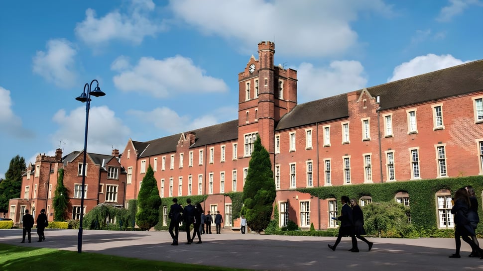 El edificio histórico con una alta torre en el campus del Trent College está rodeado de árboles y muestra a personas en camino.