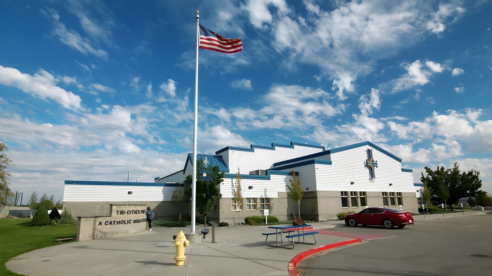 Una gran bandera americana ondea sobre el moderno edificio de la Tri-Cities Prep Catholic High School con alfombra roja y césped verde.