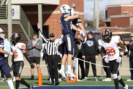 Estudiantes de la Trine University juegan un partido de fútbol americano en el campo con árbitros y edificios de ladrillo en el fondo.