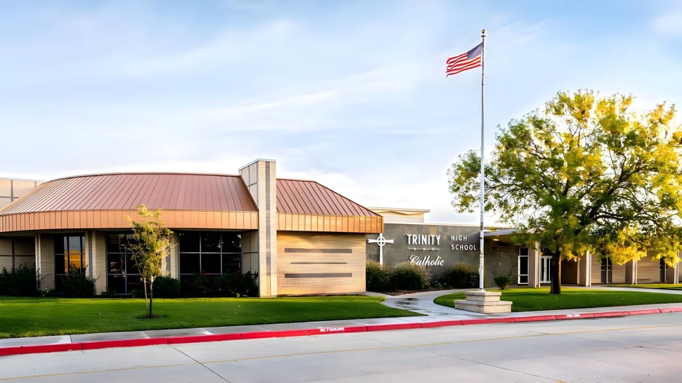 El edificio de una planta con techo de metal rojo en el terreno de la Trinity Catholic Jr/Sr High School está rodeado de césped y árboles.