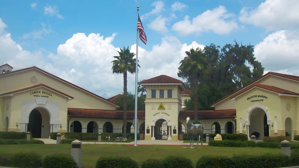 El edificio principal de la Trinity Catholic High School muestra estilo español con torre y bandera americana frente a palmeras y cielo azul.