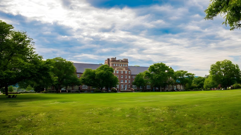 El campus de la Trinity-Pawling School muestra una gran área de césped verde frente a edificios históricos de ladrillo bajo un cielo nublado.