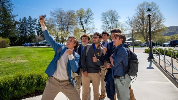 Un grupo de estudiantes está en un día soleado en el área exterior de la Trinity-Pawling School con césped y árboles de fondo.