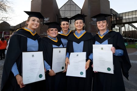 Un grupo de cinco graduadas y graduados de la University of Limerick se encuentra frente al edificio universitario sosteniendo orgullosamente sus diplomas.