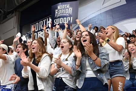 Un grupo de estudiantes celebra y anima en un evento deportivo de la University of North Florida en una arena deportiva.