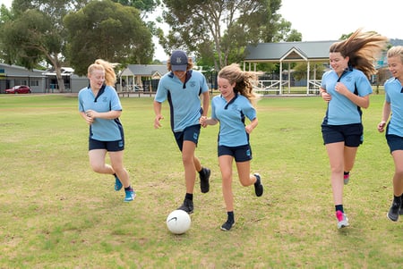 Un grupo de jóvenes atletas de la Unley High School corre juntos sobre un campo verde con el edificio escolar al fondo.