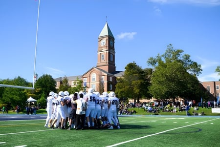 Un grupo de estudiantes en uniformes blancos está de pie sobre un campo en el terreno del Upper Canada College frente a un alto campanario.