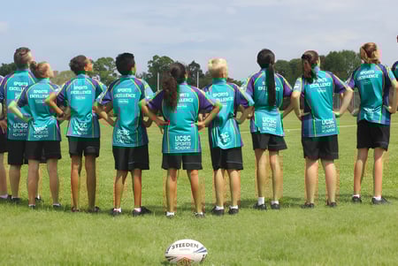 Un grupo de estudiantes del Upper Coomera State College está en un campo de fútbol junto a un balón de fútbol en primer plano.