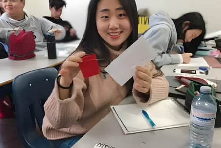 Una estudiante de la Urban International High School está sentada sonriendo en el escritorio y sostiene un sobre rojo en un aula.
