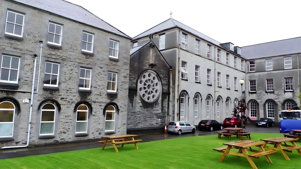 El edificio del Ursuline College Sligo con ventanas redondas y ventanas de arco rodeado de un césped con mesas de picnic.