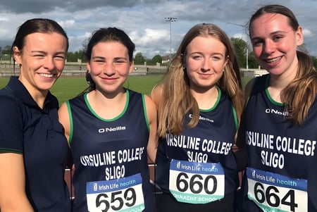 Cuatro alumnas del Ursuline College Sligo están en uniformes deportivos a juego en un campo de deportes con cielo nublado.