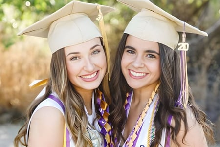 Dos estudiantes de la Valencia High School con birretes y togas posan riendo en un campo con hierba alta.