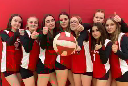 Un grupo de jóvenes estudiantes femeninas en uniforme de voleibol posan con una pelota frente a un fondo rojo en el campus del Vankleek Hill Collegiate Institute.