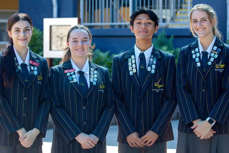 Cuatro estudiantes en ropa académica están frente a un edificio con fachada azul en el campus del Varsity College.