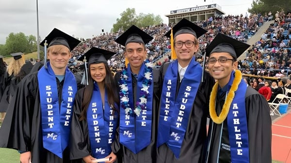 Un grupo de graduadas y graduados en togas azules y amarillas está de pie en la ceremonia de graduación del Ventura County Community College District frente a un gran público.