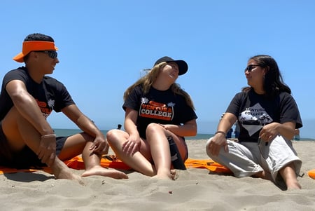 Tres personas están sentadas en la playa bajo un cielo azul en el campus del Ventura County Community College District.