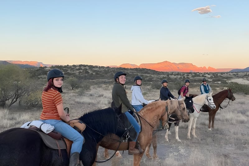 Estudiantes de la Verde Valley School montan a caballo a través de un paisaje desértico pintoresco con montañas al fondo.