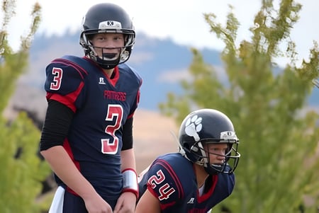 Dos jugadores de fútbol en uniforme están de pie en un campo con hierba alta frente a árboles en el terreno de la Vernon Secondary School.