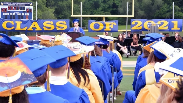 Un grupo de graduados de la Vernon Township High School está en togas azules y doradas frente a un banner que dice Class of 2021 en un campo deportivo.