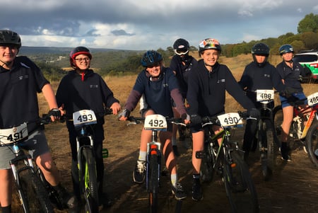 Un grupo de estudiantes de la Victor Harbor High School está en uniformes negros en una carretera de grava frente a un paisaje montañoso.