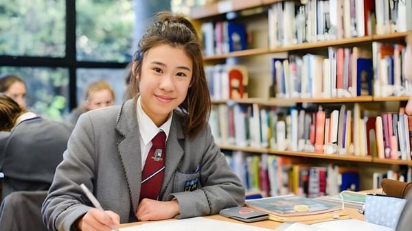 Una estudiante del Victoria College Belfast está sentada en una mesa en la biblioteca entre estanterías de libros.