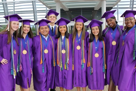 Un grupo de graduadas y graduados en togas de graduación moradas está frente a un edificio de vidrio en el campus del Virginia Beach City Public School District.