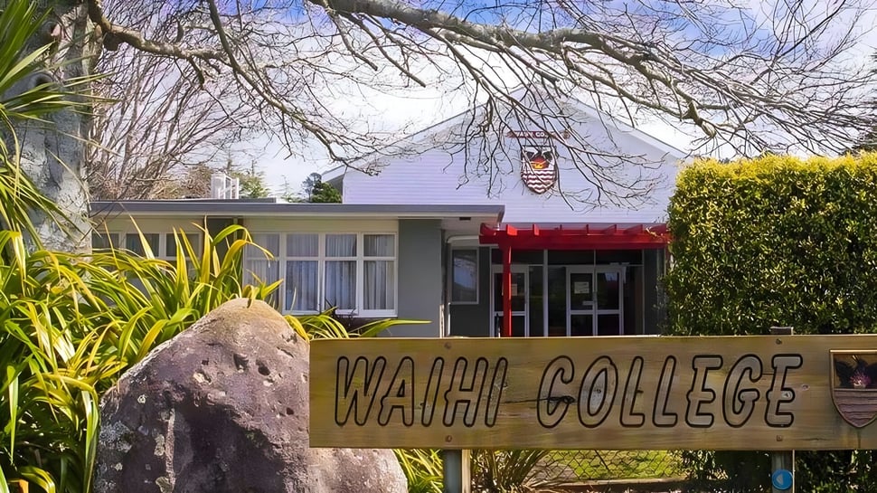 Un gran cartel con la inscripción Waihi College está rodeado de mucho verde frente a un edificio con un toldo rojo.