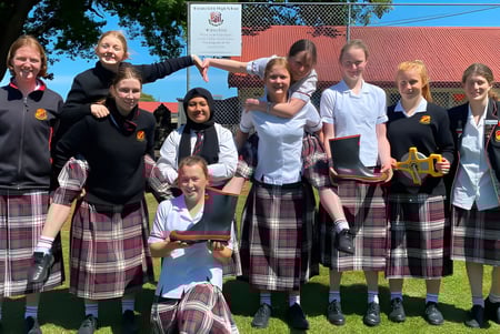 Un grupo de estudiantes de la Waitaki Girls’ High School está junto en un prado frente a un edificio con techo rojo.