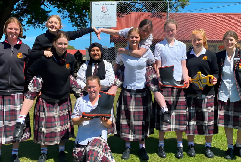 Un grupo de alumnas y alumnos de la Waitaki Girls’ High School está junto en un prado frente a un edificio de techo rojo.