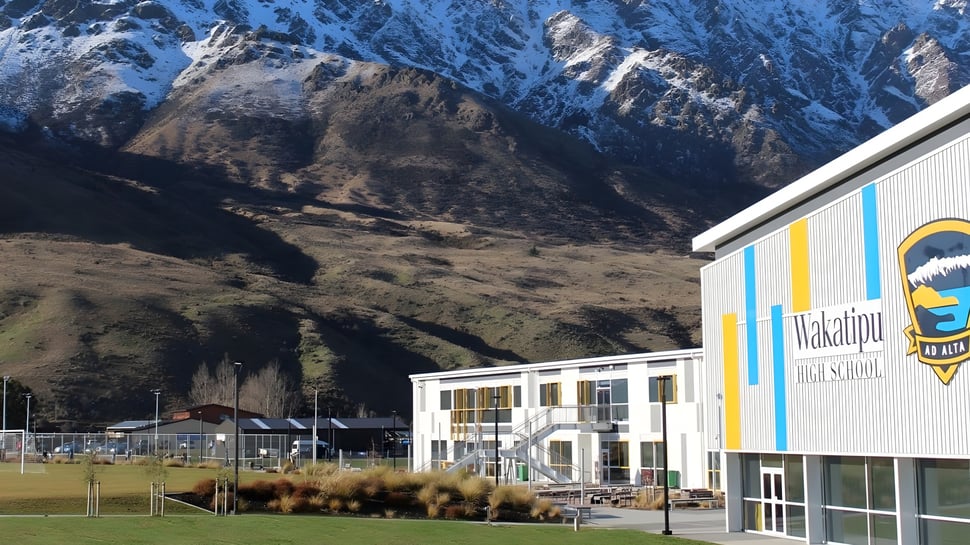 El colorido edificio de Wakatipu High School se encuentra frente a montañas cubiertas de nieve y un paisaje verde.