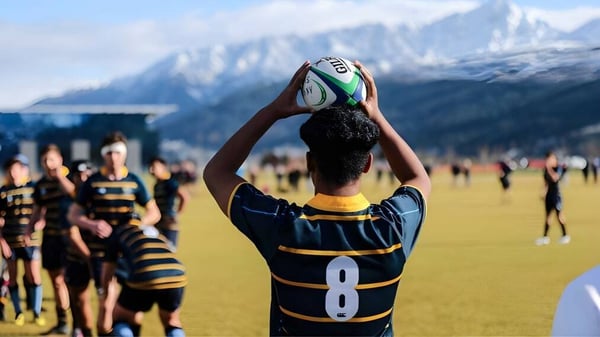 Un jugador de rugby en uniforme negro y amarillo atrapa el balón en el campo de Wakatipu High School con montañas cubiertas de nieve al fondo.