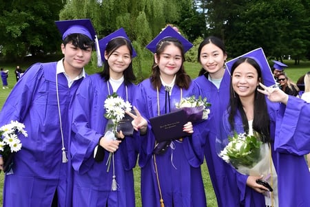 Un grupo de jóvenes mujeres de la Walnut Hill School for the Arts está en togas de graduación moradas con flores en una pradera verde.