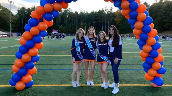 Cuatro estudiantes de la Walpole High School están juntas frente a un arco de globos en el campo de deportes.