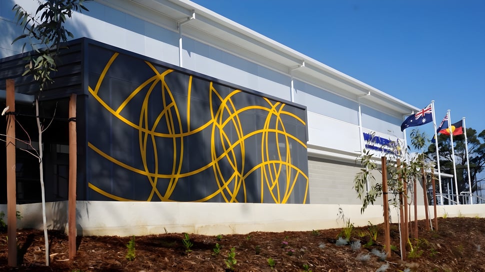 El gran edificio blanco con un mural abstracto amarillo en el campus del Wanneroo Secondary College bajo un cielo azul.