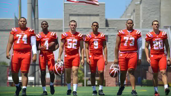 Un grupo de futbolistas de la Washington Academy está en camisetas rojas en el campo frente a un edificio con la bandera americana.