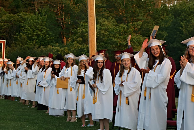 Un grupo de graduados en togas y birretes blancos está frente a un bosque en el campus de la Washington Academy.