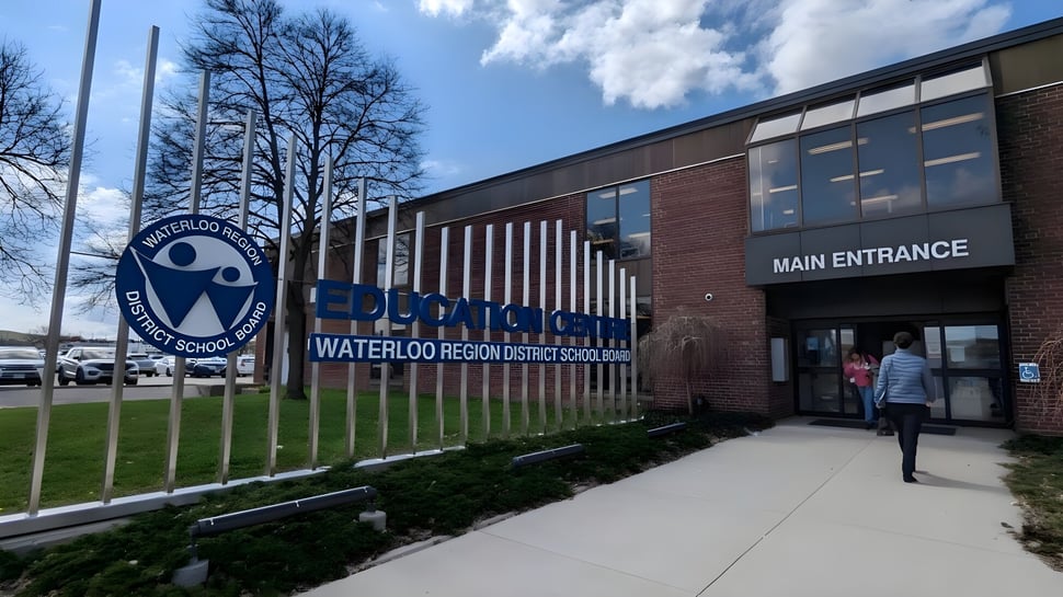 El edificio de ladrillo con la entrada y el logo del Waterloo Region District School Board está rodeado de árboles y un camino pavimentado.