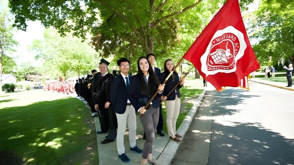 Un grupo de estudiantes de la Wayland Academy sostiene una bandera roja en un camino bordeado de árboles.