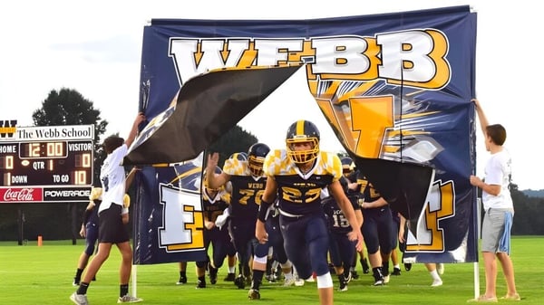 Un grupo de futbolistas entra al campo a través de un gran banner con la inscripción Webb en el campus de la Webb School.