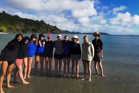 Un grupo de estudiantes del Wellington Girls' College está junto a la playa con montañas de fondo.